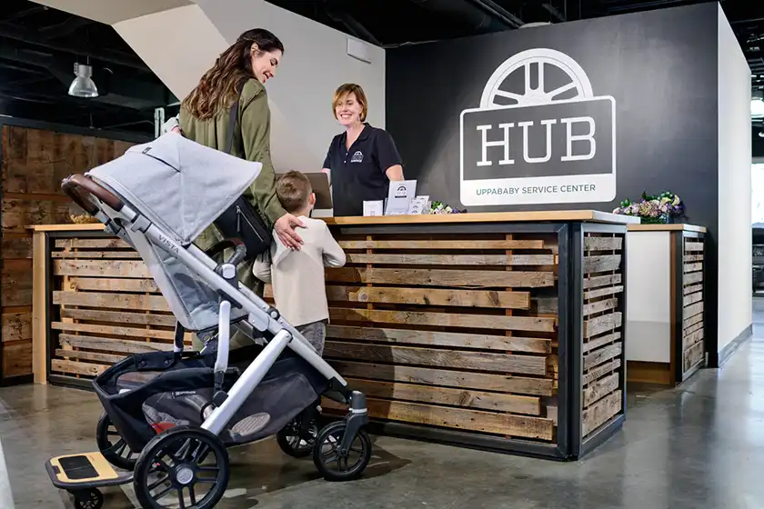 Mom and young child disembark Vista stroller and stand at wooden service counter at UPPAbaby Hub Service Center & Showroom. Staff member warmly greets mom and child, smiling as she assists them.