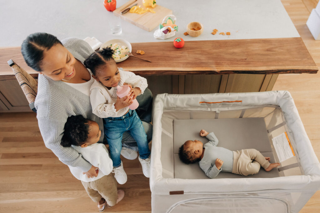 A mother with her children in an UPPAbaby Remi Playard in a family living room—Charlie—Sand Mélange.