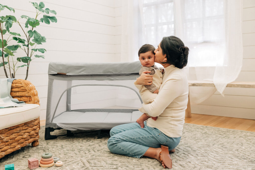 A mother holding her little one in her arms beside an UPPAbaby Remi Playard.