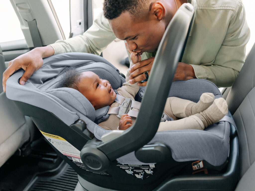 Baby laughs securely buckled into the Mesa V2 car seat as father kisses his hand. 